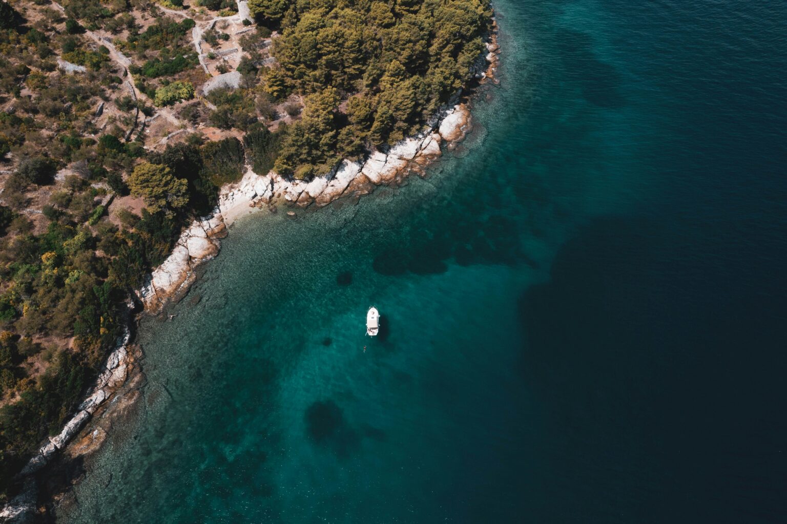 Boot in klarem türkisem Wasser an bewaldeter Küste (Luftaufnahme)